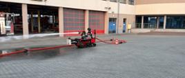 Na placu przed budynkiem JRG Centralnej Szkoły Państwowej Straży Pożarnej w Częstochowie pracuje czerwono-czarny robot gaśniczy na gąsienicach. Urządzenie podłączone jest do linii wężowej i z zamontowanej na nim prądownicy podaje silny strumień wody w kierunku otwartej przestrzeni. W tle widoczne są bramy garażowe jednostki oraz zaparkowane pojazdy pożarnicze