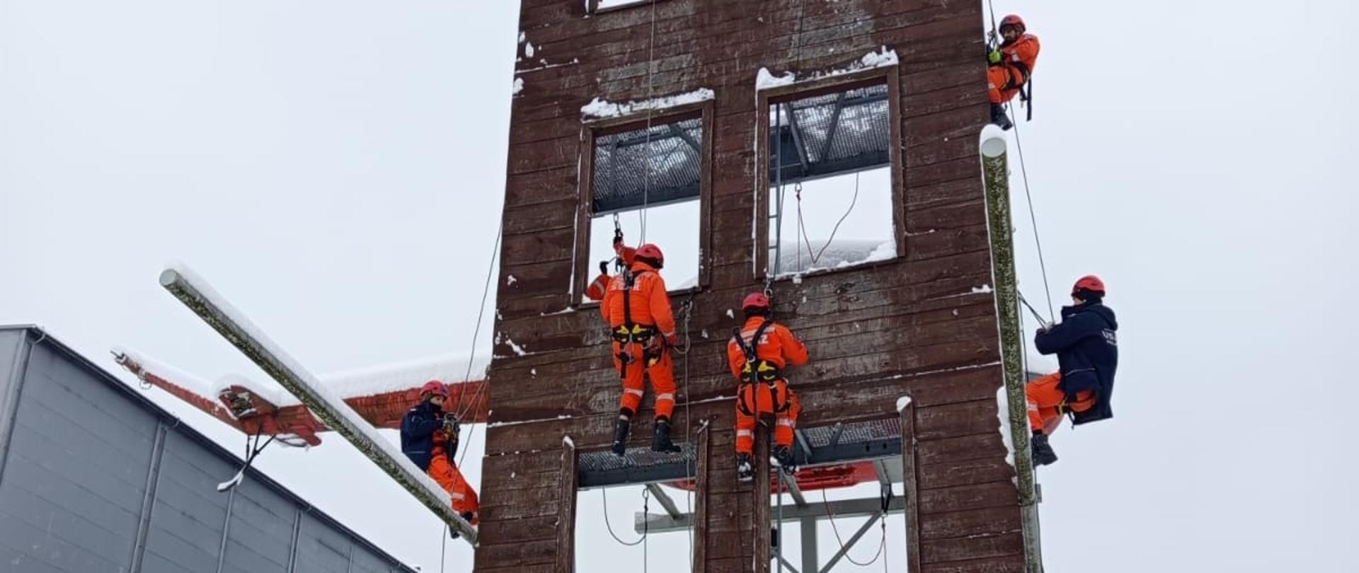 Kilku ratowników wspina się na drewnianą wieżę treningową w zimowych warunkach, używając lin i sprzętu asekuracyjnego.