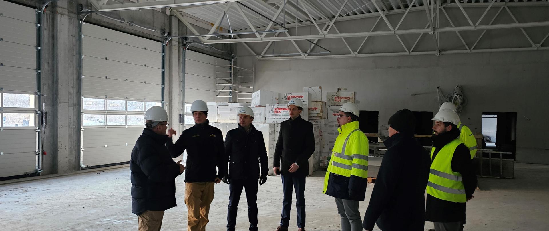 Ośmiu mężczyzn w białych kaskach na głowie w pomieszczeniu garażowym nowobudowanego obiektu podczas rozmowy.