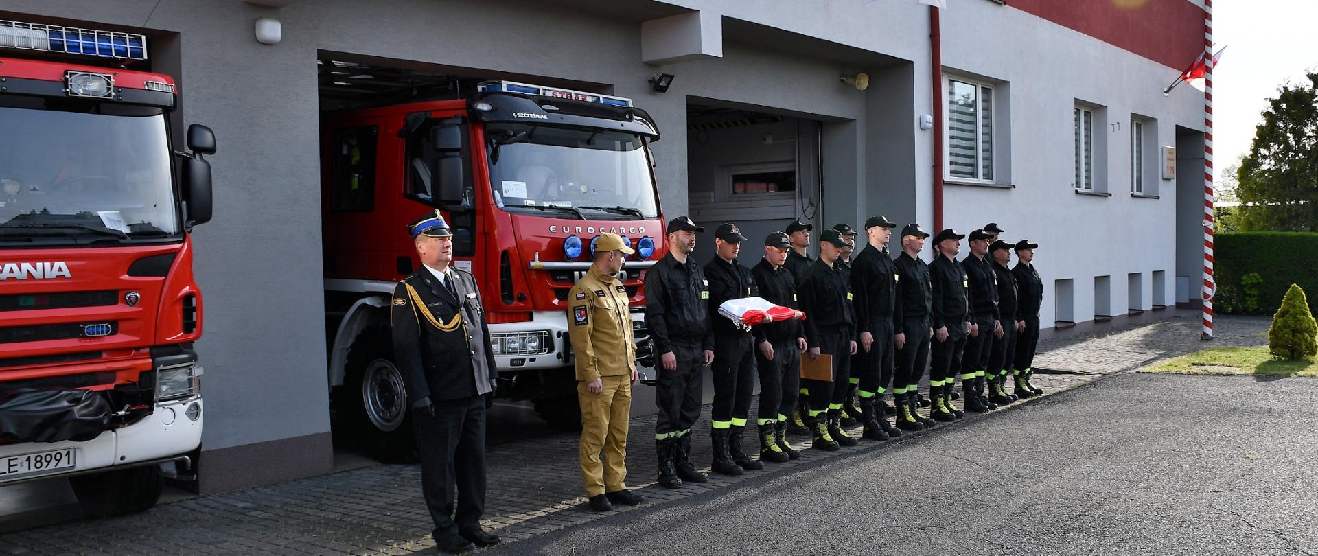Na zdjęciu widzimy komendanta oraz strażaków stojących w szeregu, jeden z nich trzyma złożoną flagę państwową.