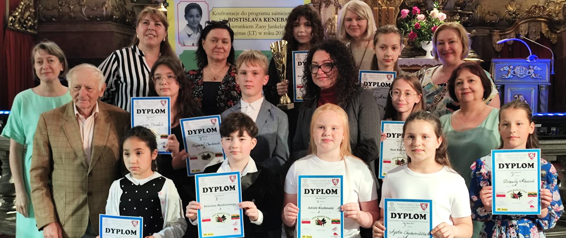 Grupowe zdjęcie uczestników i organizatorów międzynarodowego konkursu muzyki klasycznej w zabytkowym wnętrzu kościoła. Na pierwszym planie dzieci trzymają dyplomy, a jedno z nich prezentuje puchar. Za nimi stoją dorośli opiekunowie i organizatorzy. W tle widoczny jest ozdobny ołtarz oraz baner informujący o wydarzeniu „Ako-Skrzy-For”
