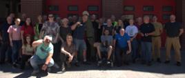 Grupa pracowników ustawiona przed budynkiem z czerwonymi bramami garażowymi, część osób stoi, część siedzi; jedno z uczestników znajduje się na wózku inwalidzkim, w tle widoczna elewacja z cegły.