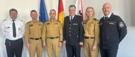 Sześć osób pozuje do wspólnego zdjęcia na tle flag Unii Europejskiej, Niemiec i Berlina. Wśród nich znajdują się przedstawiciele Berlińskiej Straży Pożarnej oraz polskich jednostek PSP. Osoby ubrane są w różne mundury – beżowe polskiej PSP oraz ciemne i białe niemieckiej straży pożarnej
