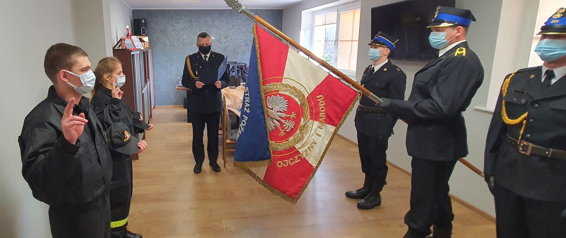 Zbiórka strażaków w jasnym pomieszczeniu. Po prawej stronie stoi trzech strażaków w mundurach wyjściowych. Środkowy trzyma biało - czerwony sztandar komendy. Po lewej dwoje funkcjonariuszy (kobieta i mężczyzna) ubranych w mundury koszarowe składa ślubowanie. W środku komendant w mundurze wyjściowym odczytuje rotę ślubowania z granatowej teczki. Wszyscy zgromadzeni mają twarze zasłonięte maseczkami. W tle wyposażenie gabinetu: szafy, stół, telewizor.
