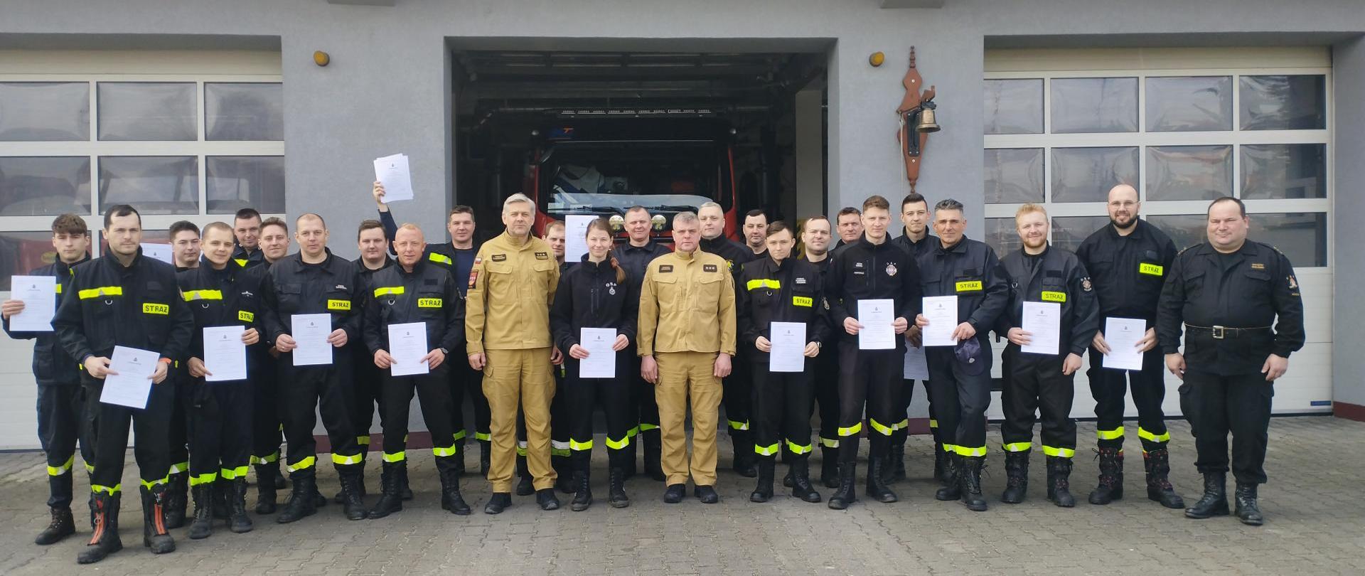 Zdjęcie przedstawia uczestników szkolenia kierowców-konserwatorów sprzętu ratowniczego OSP stojących na tle budynku komendy straży pożarnej w Koluszkach. Uczestnicy trzymają przed sobą dyplomy ukończenia szkolenia, są ubrani w czarne ubrania koszarowe, po środku w ubraniach służbowych koloru piaskowego stoją Z-ca komendanta powiatowego PSP oraz Z-ca Dowódcy JRG w Koluszkach. 