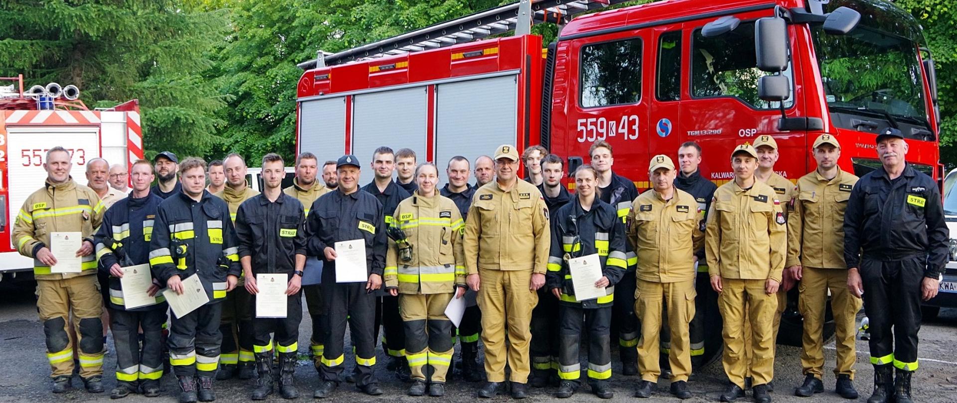 Widok ogólny z przodu. Absolwenci szkolenia i kadra PSP na tle samochodu pożarniczego.