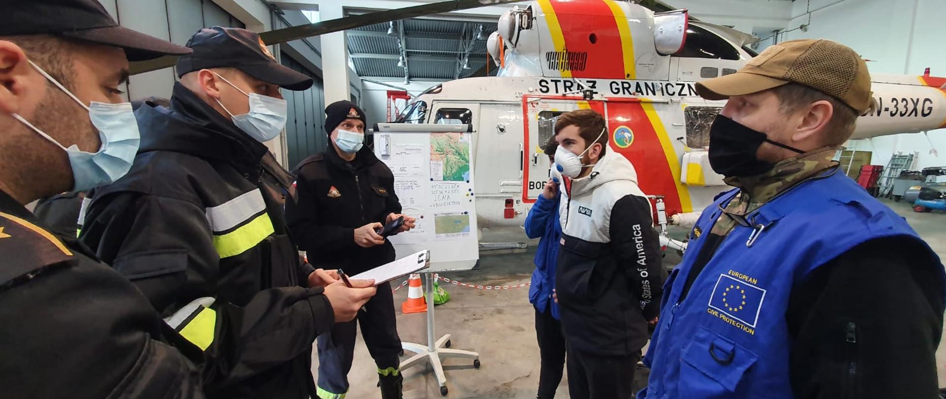 Uczestnicy szkolenia w trakcie zajęć praktycznych. Trzech strażaków, cywil i dwóch przedstawicieli UE w trakcie wymiany informacji. W tle śmigłowiec straży granicznej. 