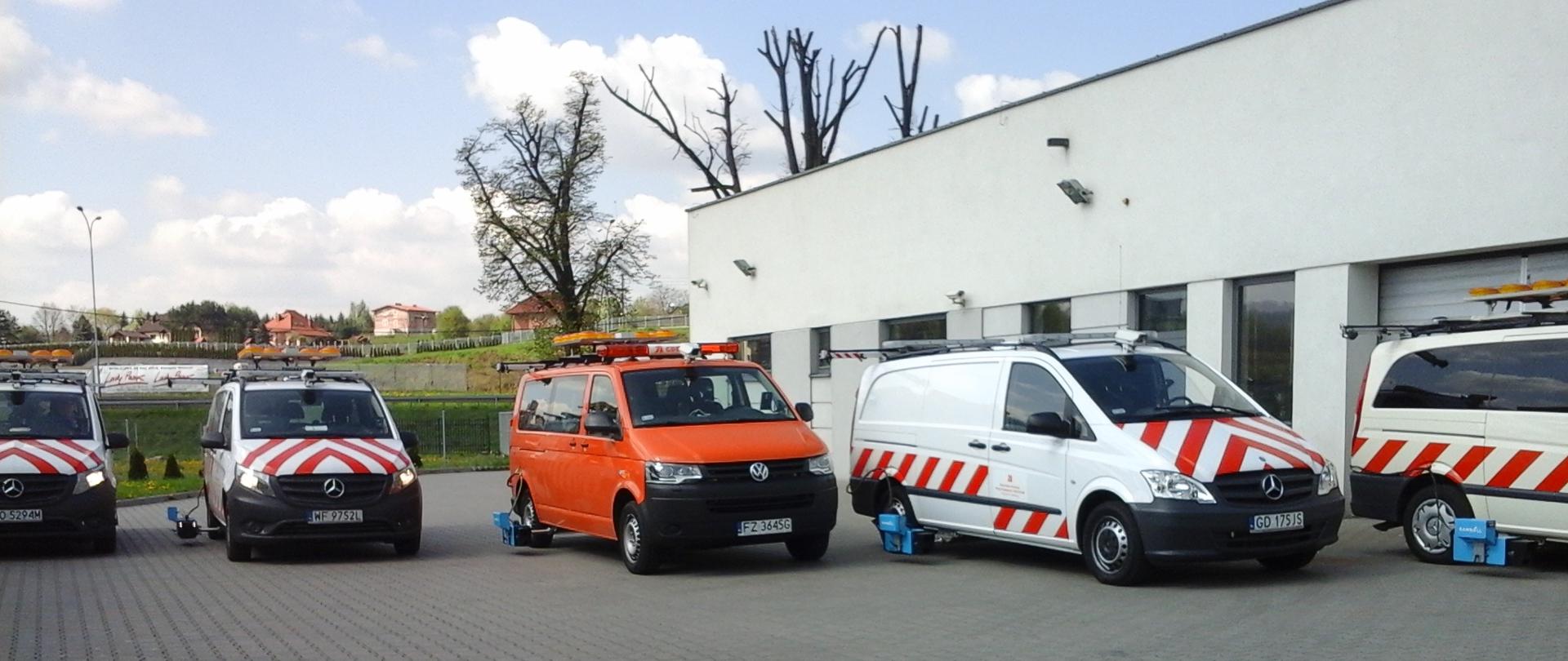 Cztery samochody techniczne zaparkowane na placu przed budynkiem. Pojazdy w kolorach białym i pomarańczowym, z pasami ostrzegawczymi oraz sprzętem pomiarowym zamontowanym z tyłu. W tle budynek użytkowy i zieleń.
