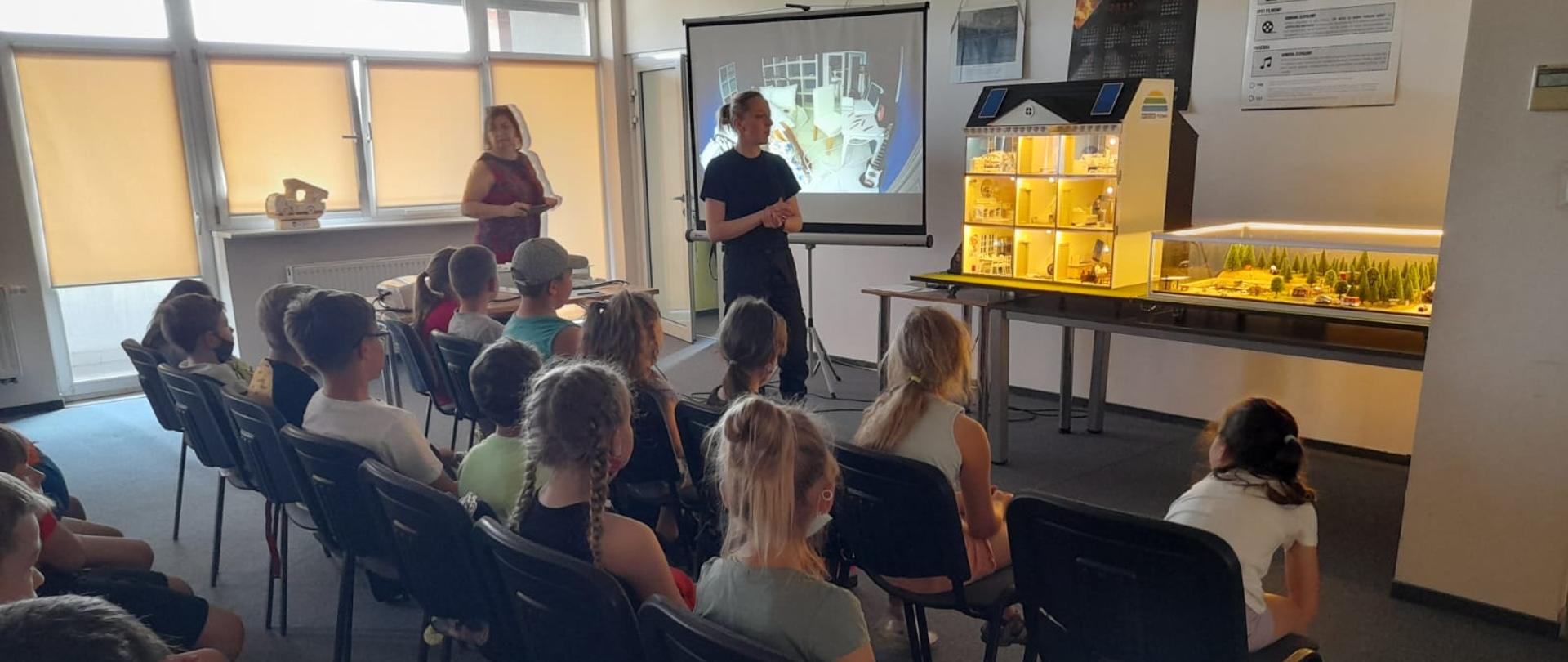 Sala edukacyjna. Dzieci siedzą na krzesłach i słuchają funkcjonariuszkę straży pożarnej. Przed strażakiem mobilne symulatory zagrożeń: dom oraz las.