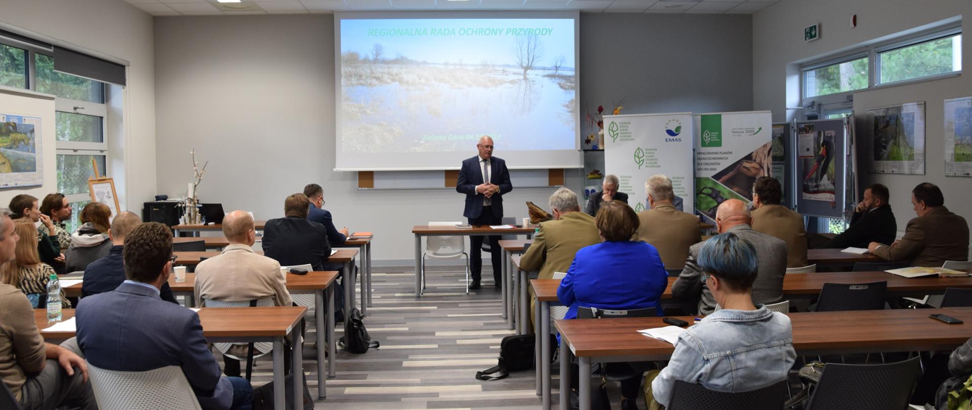 W dużej sali za stołami siedzi grupa kilkudziesięciu kobiet i mężczyzn. Przed nimi za stołem siedzi dwóch mężczyzn, jeden ubrany w garnitur stoi i przemawia.