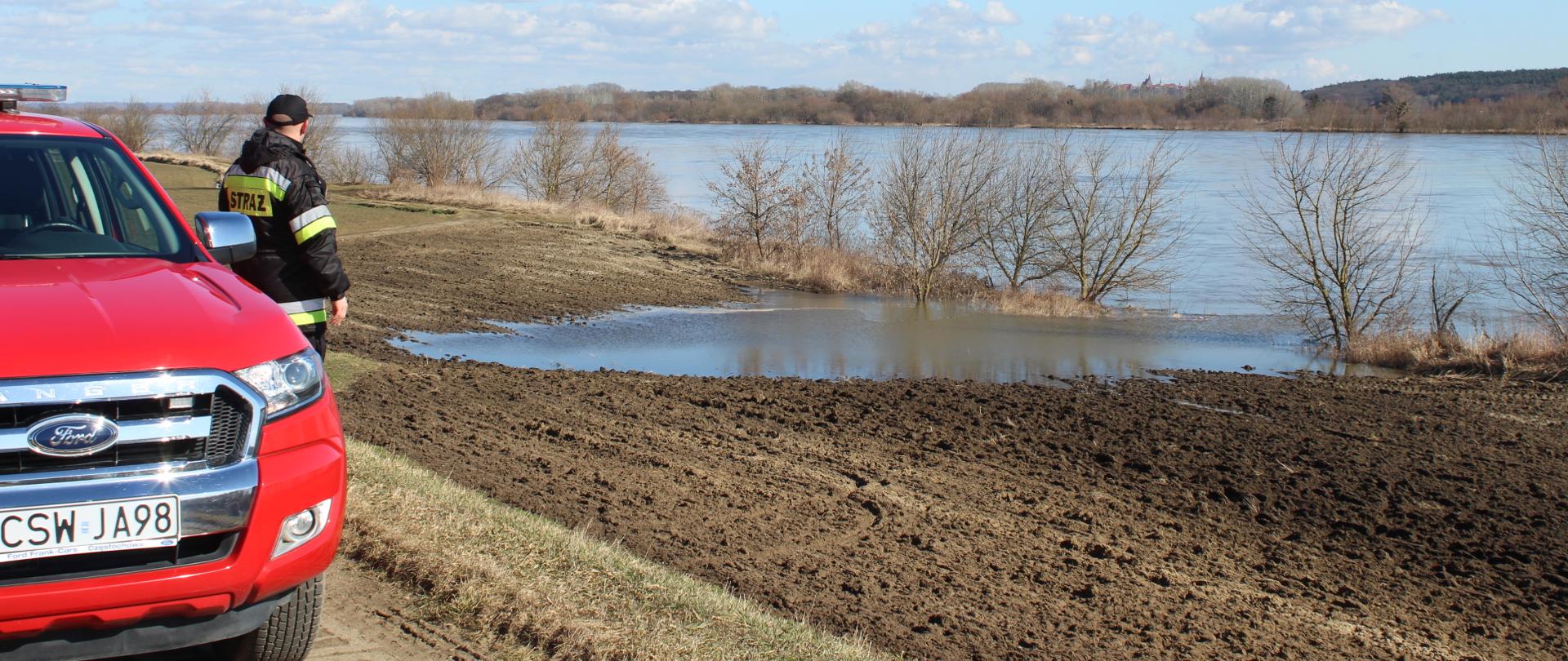 Na wale przeciwpowodziowym po lewej stronie widać przód samochodu operacyjnego straży pożarnej przy którym stoi młodszy brygadier Paweł Puchowski i obserwuje poziom wody w Wiśle 