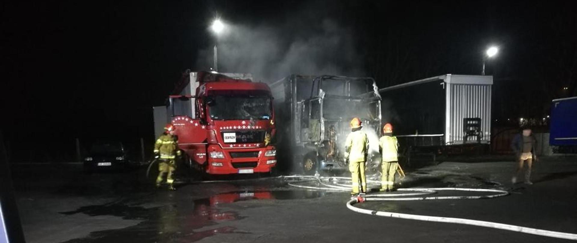 Na placu zaparkowane są dwa ciągniki siodłowe z naczepami. Strażacy dogaszają wodą z linii wężowych pojazdy, które uległy spaleniu.