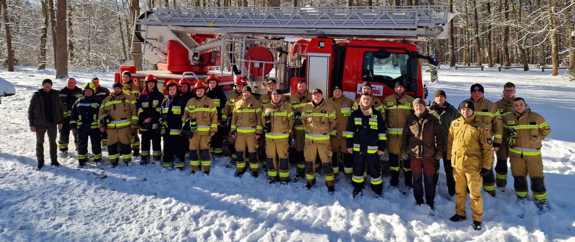 Zdjęcie grupowe strażaków biorących udział w ćwiczeniach. Stoją na tle wozu strażackiego oraz lasu