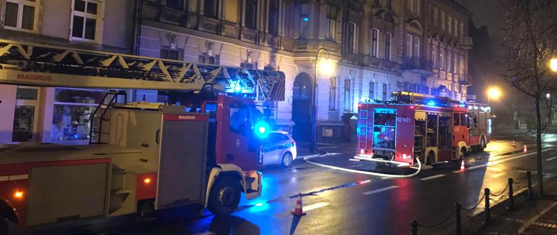 Jest pora nocna. Na zdjęciu widać trzy samochody pożarnicze stojące na jezdni przed ciągiem kamienic. Jeden pas ruchu jest wygrodzony pachołkami ostrzegawczymi a z środkowego samochodu pożarniczego rozciągnięta jest linia gaśnicza