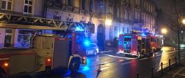 Jest pora nocna. Na zdjęciu widać trzy samochody pożarnicze stojące na jezdni przed ciągiem kamienic. Jeden pas ruchu jest wygrodzony pachołkami ostrzegawczymi a z środkowego samochodu pożarniczego rozciągnięta jest linia gaśnicza
