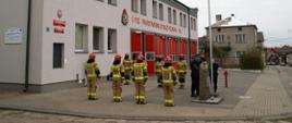 Na zdjęciu widać strażaków w ubraniach bojowych, stojących w dwóch rzędach przed budynkiem komendy. Na pierwszym planie poczet flagowy dokonuje podniesienia flagi na maszt.