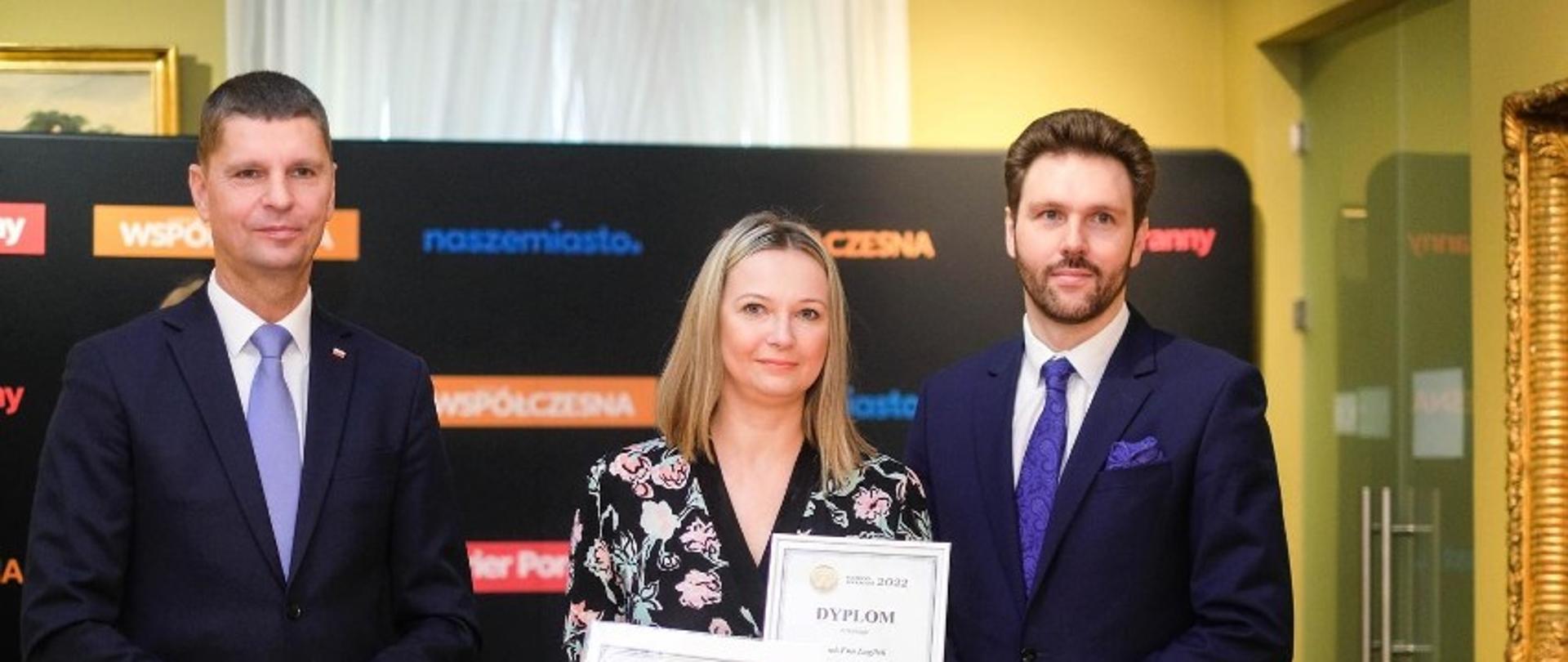 Wiceminister Piontkowski stoi obok ubranej w suknię w kwiaty kobiety trzymającej w rękach dwa dyplomy i mężczyzny w granatowym garniturze.