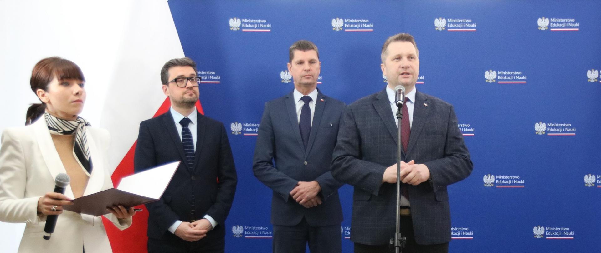 Na tle niebieskiej ściany stoi trzech mężczyzn w garniturach i kobieta w białej marynarce, trzymająca w ręku bezprzewodowy mikrofon. Za nimi z boku polska flaga.