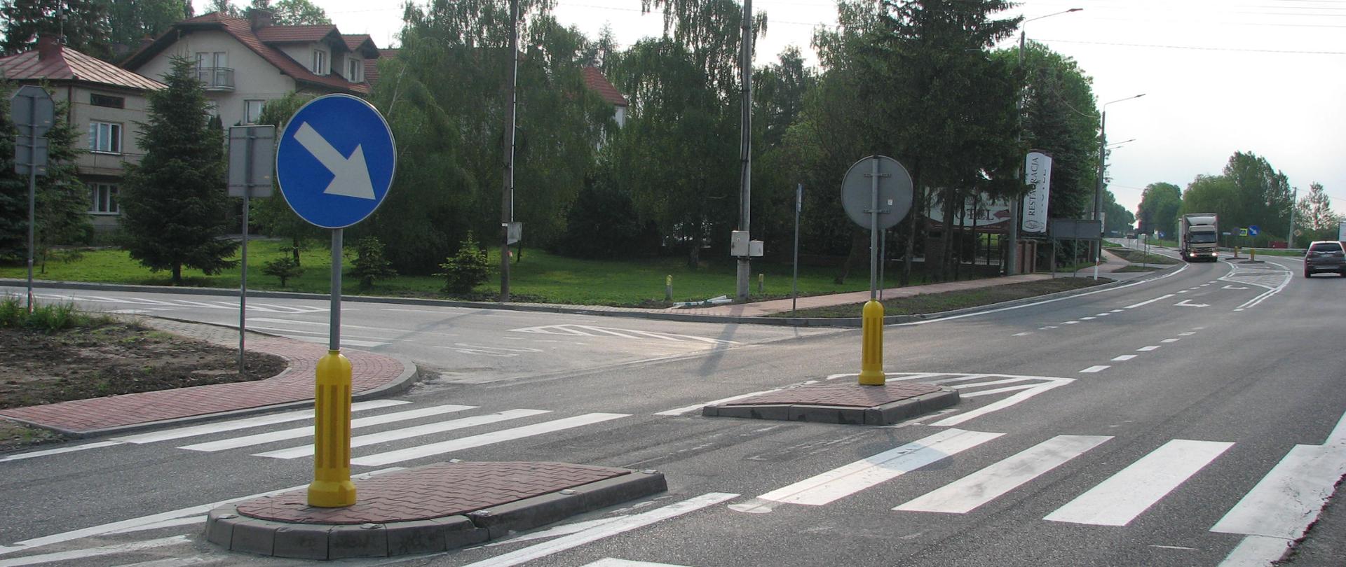 DK73 Stopnica - skrzyżowanie z przejściem i azylem dla pieszych. Na pierwszym planie na azylu niebieski znak drogowy z ukośną białą strzałką - nakaz jazdy po prawej stronie znaku. Po lewej stronie dom jednorodzinny. W oddali na drodze samochody. 