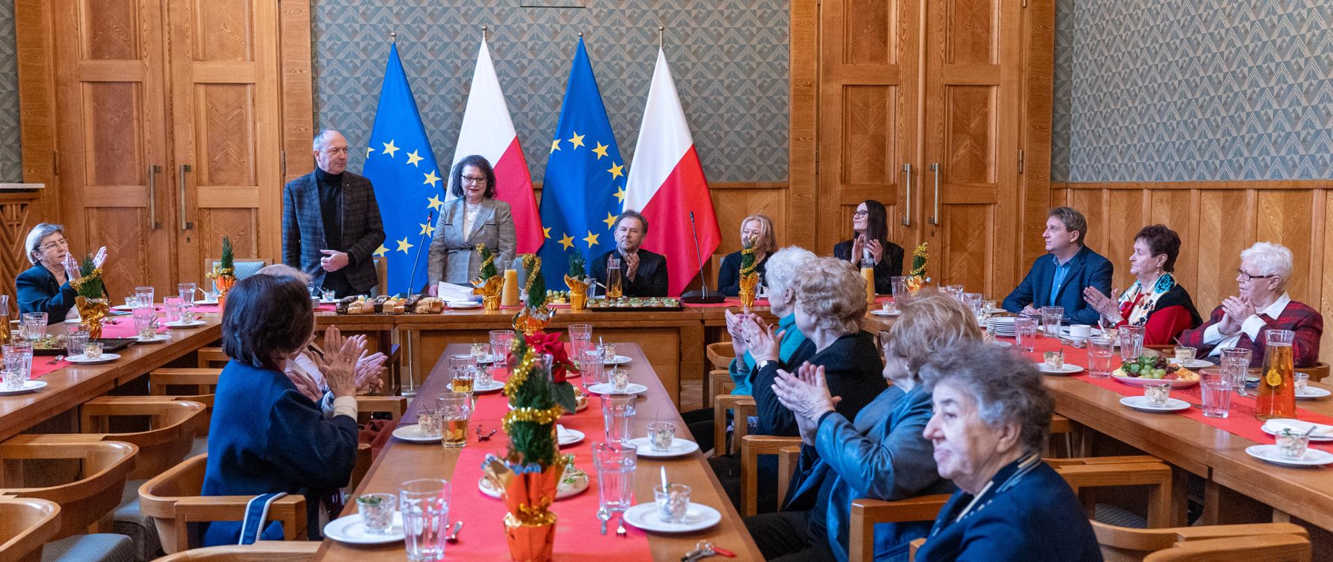 Widok na salę konferencyjną z długimi stołami nakrytymi czerwonymi bieżnikami. Na środku, pod flagami Polski i Unii Europejskiej, stoją przemawiające osoby. Seniorzy siedzący przy stołach klaszczą.