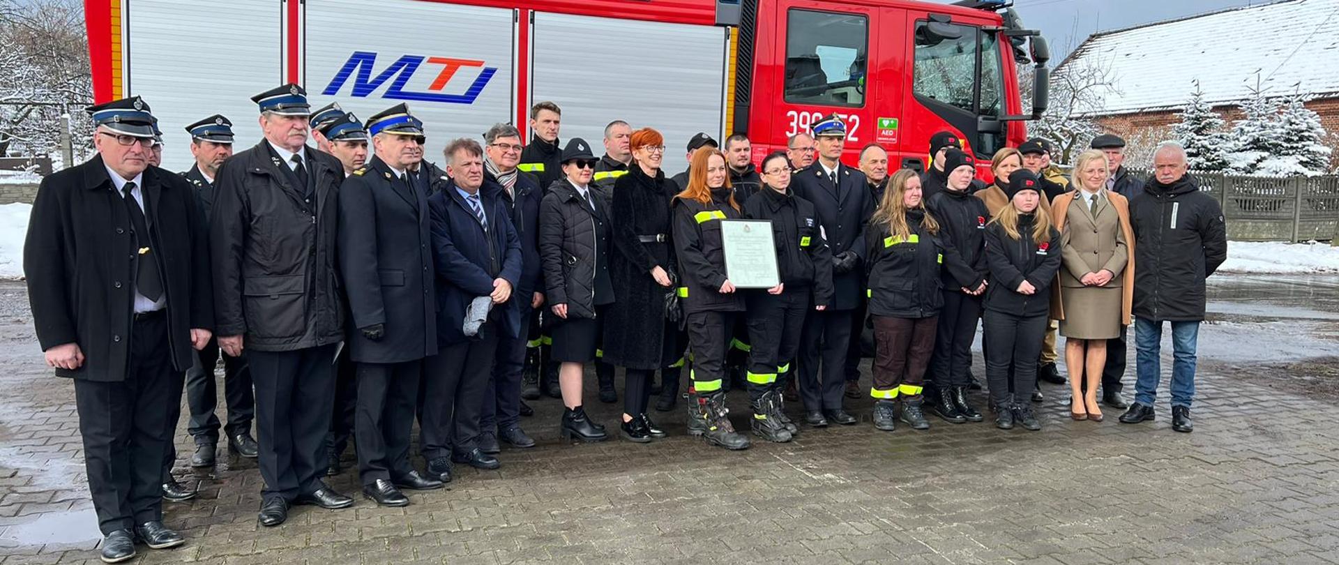 Umundurowani strażacy PSP i OSP wraz z zaproszonymi gośćmi stojący na tle samochodu gaśniczego.
