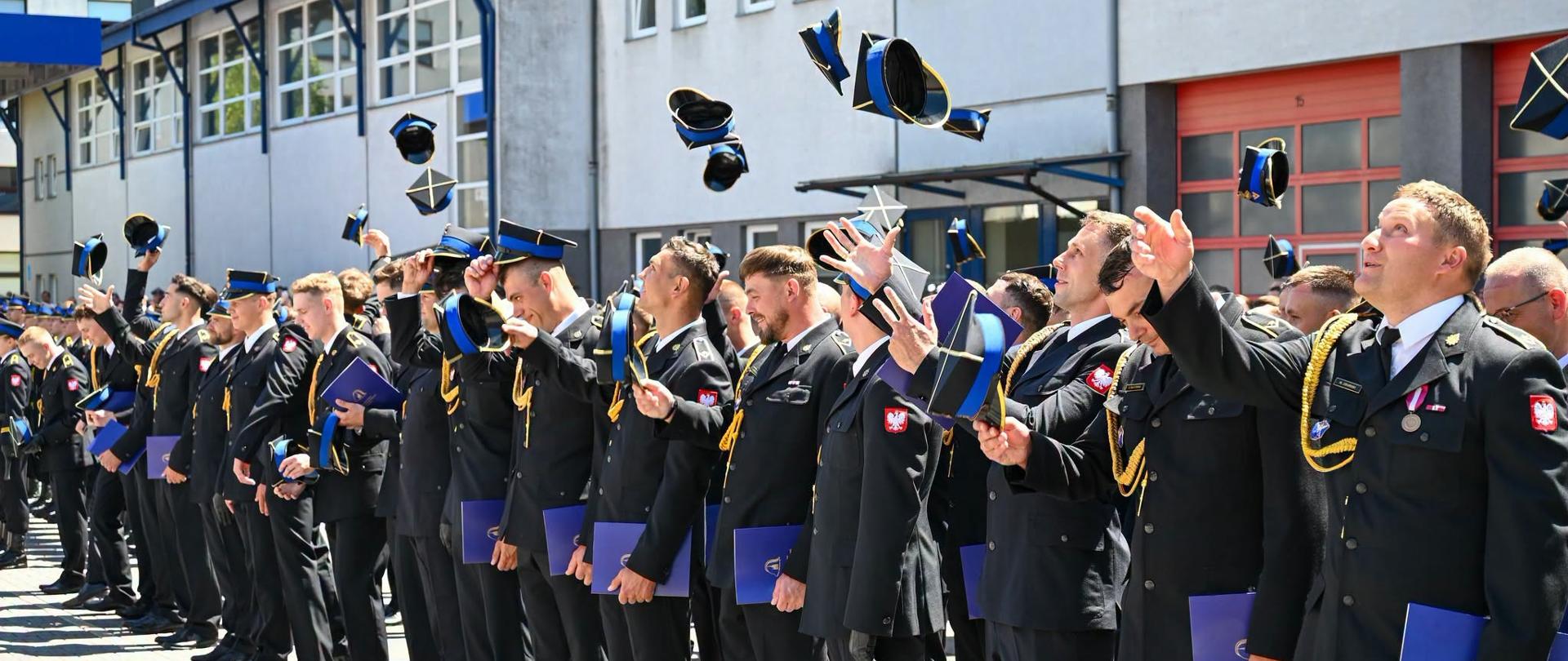 Grupa strażaków Państwowej Straży Pożarnej w galowych mundurach stoi w szeregu na placu przed budynkiem komendy. Wszyscy z uśmiechem i radością podrzucają w górę swoje czapki rogatywki podczas uroczystego apelu. W rękach trzymają niebieskie foldery lub dyplomy. W prawym dolnym rogu widnieje podpis: „st. str. kdt Piotr Krzak | Państwowa Straż Pożarna”
