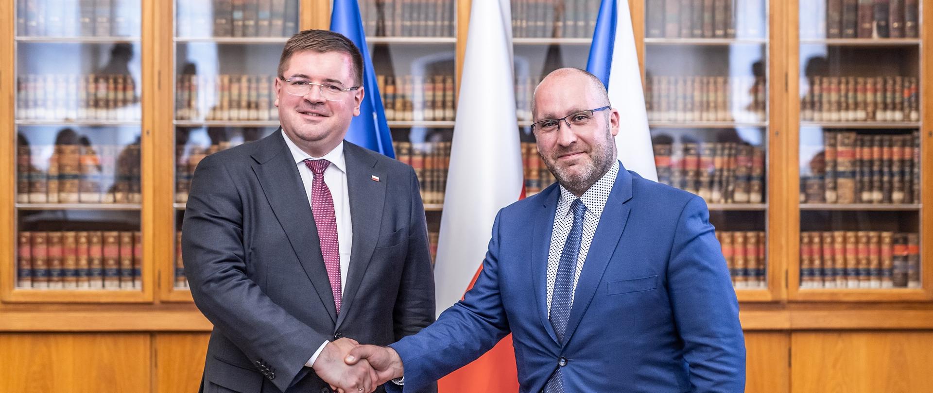 Wiceminister Rzymkowski stoi obok mężczyzny w garniturze, podają sobie ręce, za nimi polska i czeska flaga, dalej szafy z książkami.
