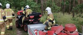 Quad z przyczepą na której znajdują się zwinięte węże, z boku strażacy, sceneria leśna.