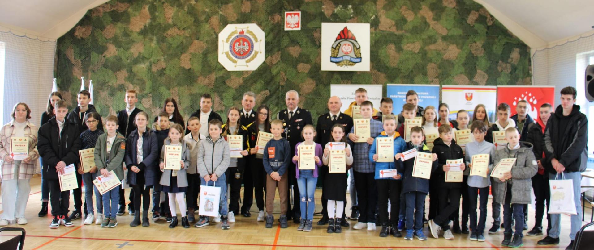 Zdjęcie ukazuje dzieci zgromadzone na sali gimnastycznej stojące i trzymające w rękach dyplomy i nagrody za uczestnictwo w etapie powiatowym Ogólnopolskiego Turnieju Wiedzy Pożarniczej. Z tyłu stoją przedstawiciele PSP i OSP .