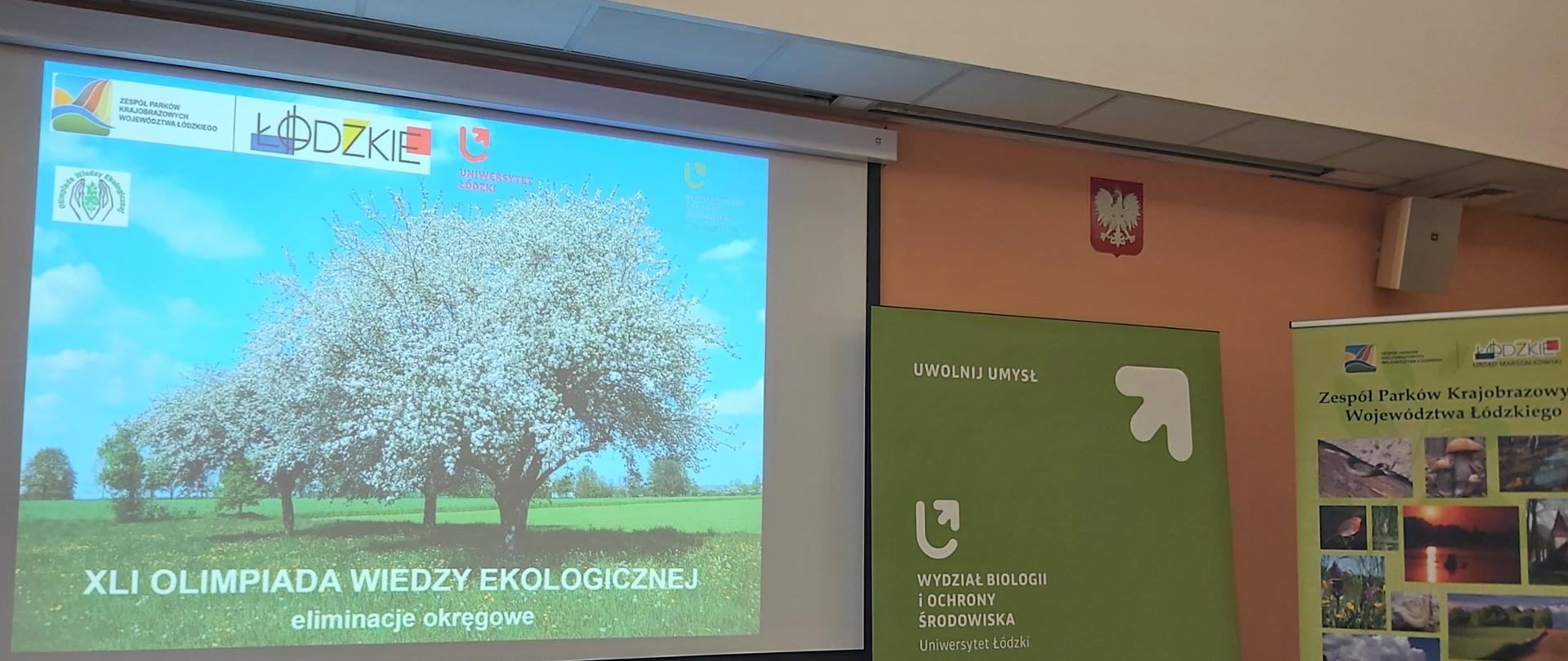 Sala konferencyjna z projektorem. Na ekranie wyświetlony slajd z kwitnącym drzewem na łące i napisem o 41. Olimpiadzie Wiedzy Ekologicznej – eliminacjach okręgowych. Obok stoją roll-upy promujące Wydział Biologii i Ochrony Środowiska Uniwersytetu Łódzkiego oraz Zespół Parków Krajobrazowych Województwa Łódzkiego. W sali widać stół i krzesła