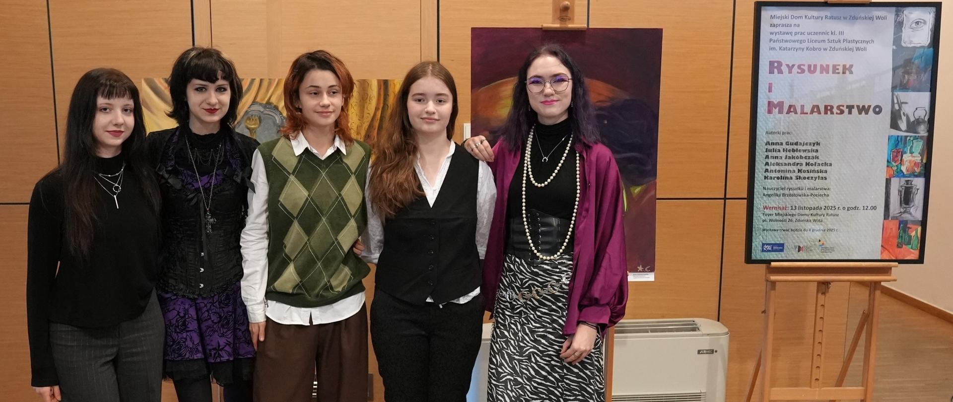 Zdjęcie: pięć dziewczyn, stojących przy sztaludze z plakatem informującym o wystawie prac rysunku i malarstwa.