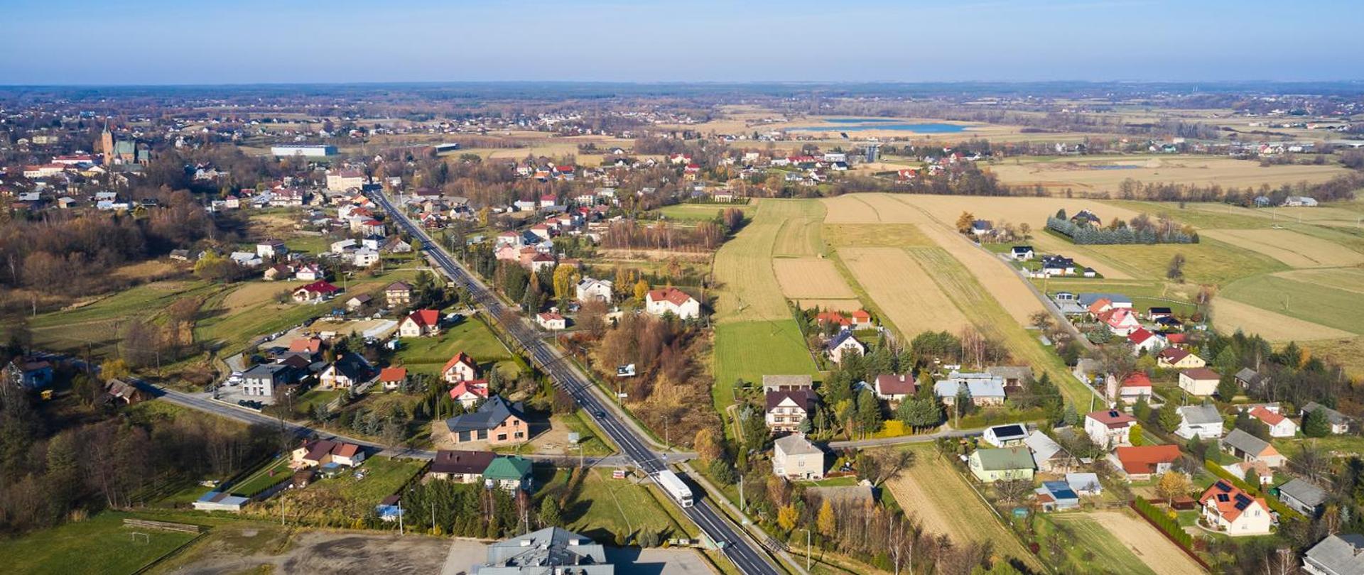 Zdjęcie z lotu ptaka pokazuje niewielką miejscowość z domami jednorodzinnymi i polami uprawnymi. Przez środek biegnie prosta droga z kilkoma pojazdami. W tle widoczne są kolejne zabudowania, zbiornik wodny oraz wieża kościoła. Całość oświetla słoneczny, bezchmurny dzień.
