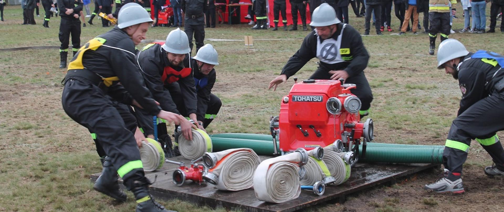 Fotografia przedstawia drużynę męską grupy A podczas wykonywania zadania ćwiczenia bojowego. Zawodnicy ubrani w umundurowanie bojowe z ubranymi pasami strażackimi. Każdy z zawodników posiada oznakowanie przewidziane regulaminem zawodów. Na głowach ubrane mają hełmy. Pięciu zawodników zabiera sprzęt z podestu podczas wykonywania swoich zadań w konkurencji. W tle zdjęcia licznie zgromadzona publiczność na stadionie dopinguje drużynę.