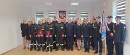 Grupowe zdjęcie w sali uroczystości – na środku stoją młodzi strażacy w mundurach koszarowych z dyplomami w rękach, obok nich oficerowie w galowych mundurach PSP oraz poczet sztandarowy z flagą jednostki. W tle godło Polski na ścianie.