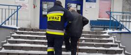 Zdjęcie przedstawia strażaka w ubraniu bojowym pomagającemu starszemu mężczyźnie wejście po schodach do budynku w celu szczepienia przeciwko koronawirusowi