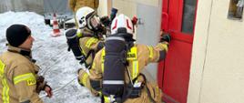 Podczas egzaminu praktycznego rota strażaków w aparatach powietrznych przed drzwiami do budynku. W pobliżu dwóch strażaków nadzorujących ćwiczenie.