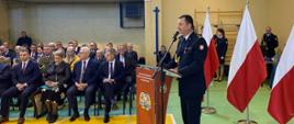 Komendant Powiatowy PSP w Ciechanowie podczas przemówienia. Stoi przy mównicy, za nim 3 flagi biało-czerwone. W oddali widać zaproszonych gości oraz funkcjonariuszy KP PSP w Ciechanowie.