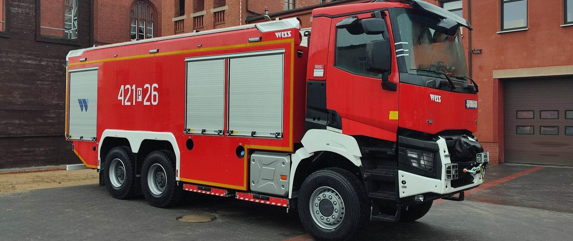 Czerwony samochód pożarniczy ze srebrnymi żaluzjami skrytek na sprzęt stojący placu z szarej kostki brukowej. W tle budynki