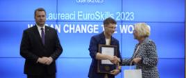 Minister Czarnek, wiceminister Machałek i młody człowiek w granatowej marynarce, z medalem na szyi stoją przed wielkim ekranem z napisem Laureaci EuroSkills 2023.