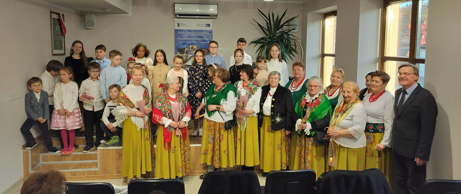 Wnętrze auli Państwowej Szkoły Muzycznej I i II stopnia w Nowym Targu, filia w Jaworkach. W głębi, na podwyższeniu widoczni uczniowie szkoły biorący udział w koncercie z okazji Dnia Kobiet jako wykonawcy. Z przodu, przed podwyższeniem zaproszone Panie z Koła Gospodyń Wiejskich w Jaworkach w strojach regionalnych z wręczonymi kwiatami. Z prawej strony kierownik szkoły. W dole krzesła oraz widownia.