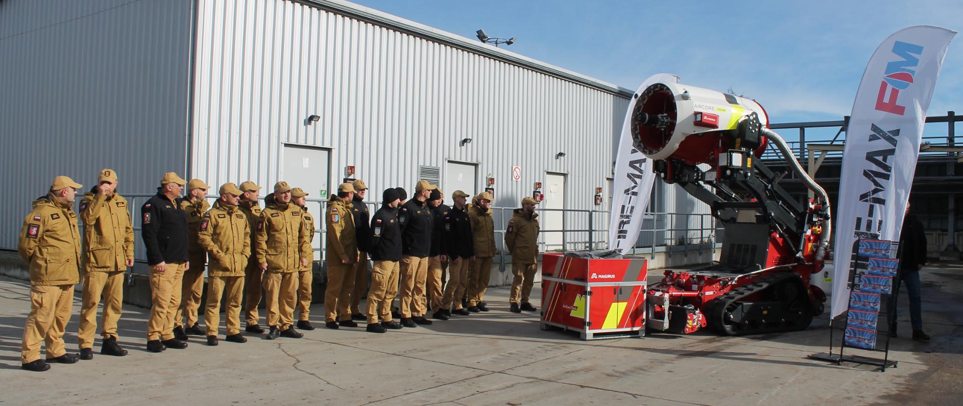 Ujęcie prezentacji lub szkolenia w obiekcie przemysłowym. Robot gaśniczy Magirus AirCore TAF60, z widocznym logo i dmuchawą, stoi na pierwszym planie obok standu z reklamą. Przed robotem stoi szereg kilkunastu strażaków w mundurach koszarowych w kolorze piaskowym i czarnych polarach, ustawionych w szyku na betonowym placu. Tło stanowi duży, biały budynek magazynowy z metalowej blachy.