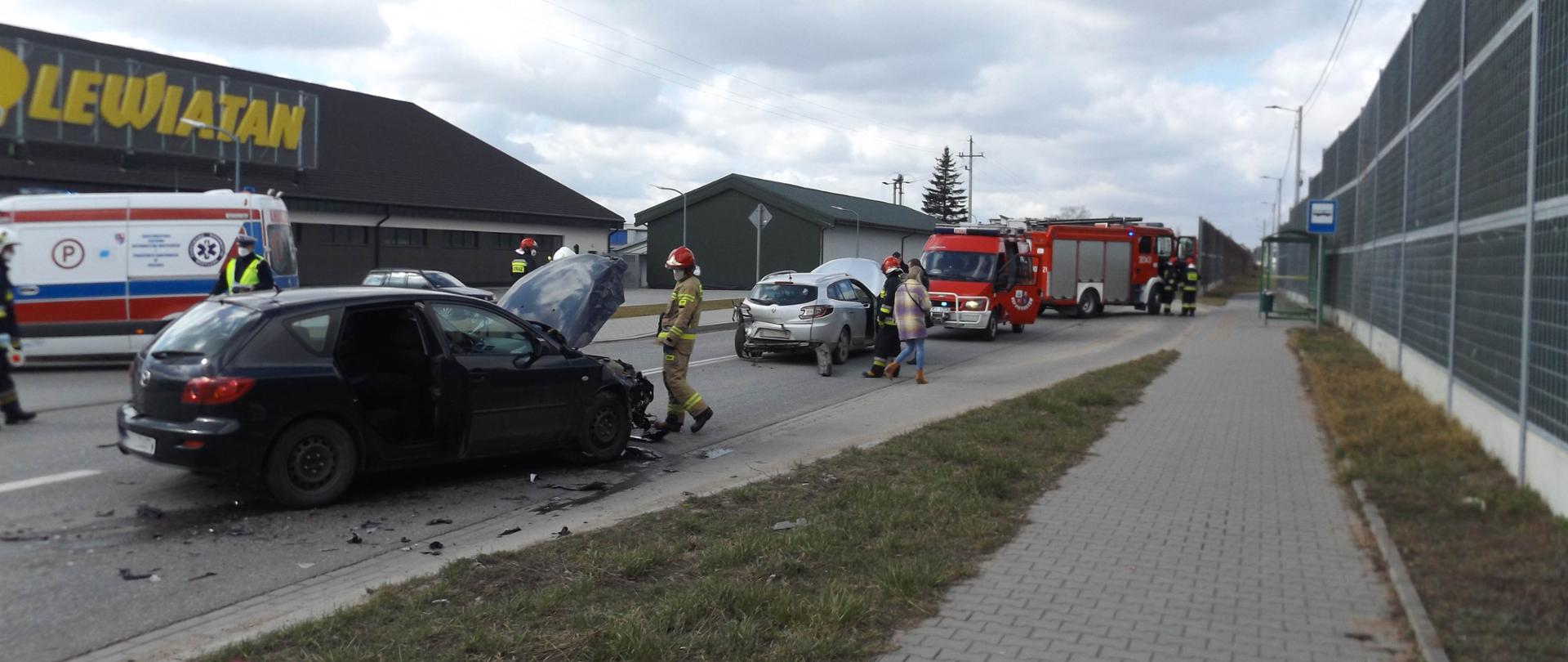 Zdjęcie przedstawia wypadek samochodowy – czarna mazda kombi z podniesioną maską, obok niej połamane plastiki i inne części. Przy samochodzie stoi strażak. Dalej widać uszkodzone srebrne Renault i samochody Straży Pożarnej. W tle stoi Pogotowie Ratunkowe.