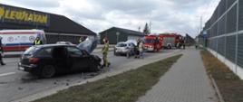 Zdjęcie przedstawia wypadek samochodowy – czarna mazda kombi z podniesioną maską, obok niej połamane plastiki i inne części. Przy samochodzie stoi strażak. Dalej widać uszkodzone srebrne Renault i samochody Straży Pożarnej. W tle stoi Pogotowie Ratunkowe.