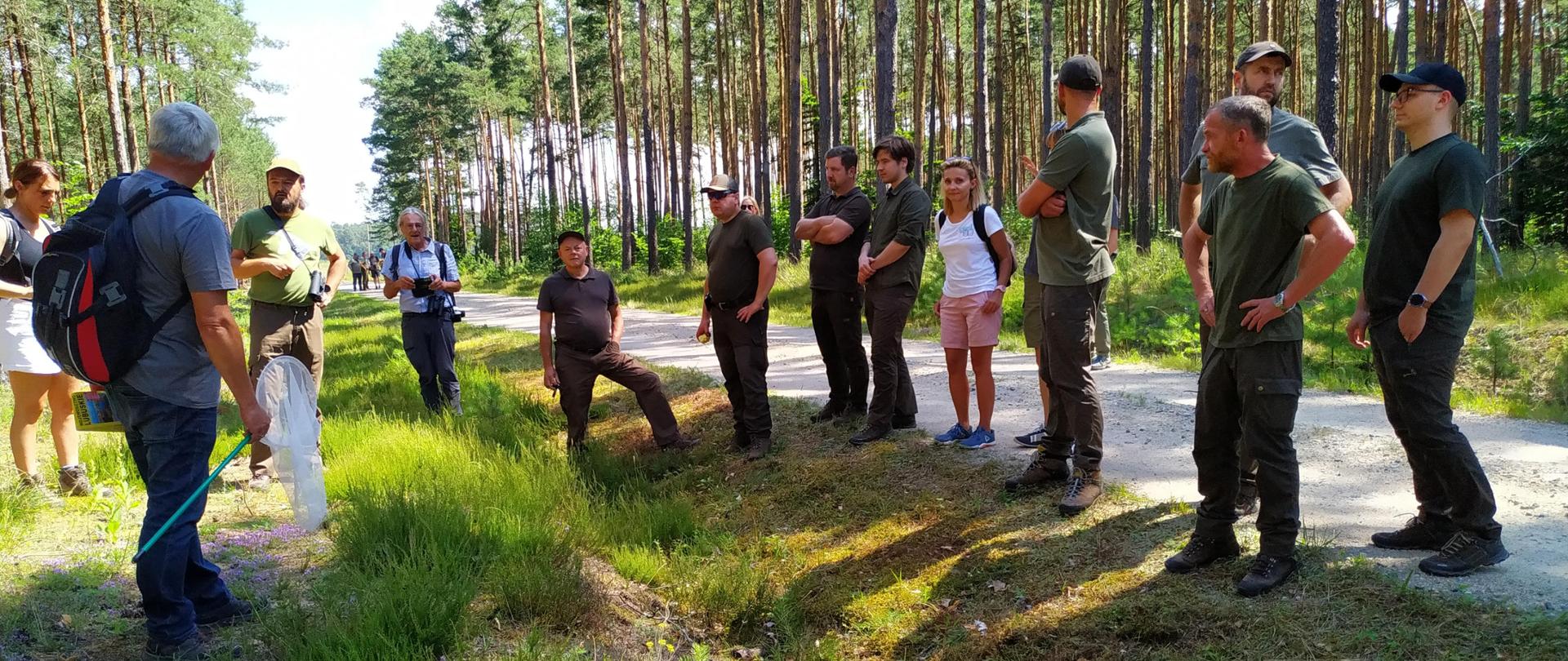 Grupa kilkunastu osób stoi na leśnej ścieżce. Na drugim planie pnie drzew iglastych- sosny.