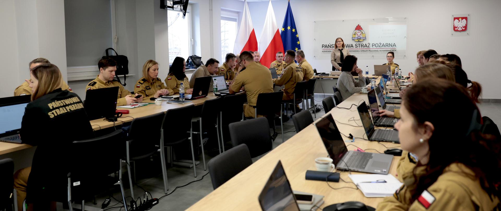 zdjęcie przedstawia salę szkoleniową Komendy Wojewódzkiej straży pożarnej w Katowicach podczas szkolenia dotyczącego ochrony danych osobowych. W pomieszczeniu ustawione są długie, jasne stoły w kształcie litery U. Przy stołach siedzi około dwudziestu osób - strażaków i pracowników cywilnych. Większość z nich ma na sobie piaskowe mundury służbowe z naszywką polskiej flagi na ramieniu. Kilka osób jest ubranych cywilnie. Uczestnicy siedzą na krzesłach i pracują przy otwartych laptopach. Na stołach znajdują się także notesy, długopisy, kubki z kawą, butelki z wodą oraz ładowarki i przewody. Niektórzy patrzą w ekrany komputerów, inni rozmawiają z osobami obok lub słuchają prowadzącej. Na końcu sali stoi kobieta prowadząca szkolenie. Za nią znajdują się trzy flagi: Polski, Polski oraz Unii Europejskiej. Na ścianie widoczna jest tablica z napisem „Państwowa Straż Pożarna” oraz godło Polski – biały orzeł na czerwonym tle. Sala jest jasna. Ma białe ściany, duże okna z roletami oraz sufit z panelowym oświetleniem. Pod sufitem zamontowany jest projektor, który służy do wyświetlania prezentacji.