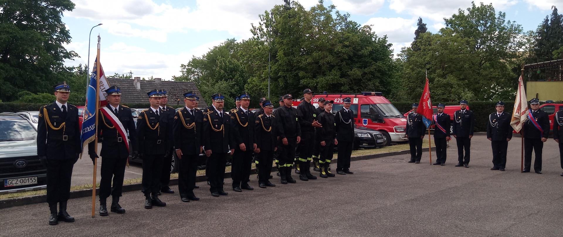 Poczty sztandarowe oraz strażacy, którzy stoją w dwuszeregu na placu Komendy Powiatowej PSP w Człuchowie. W tle samochody pożarnicze.