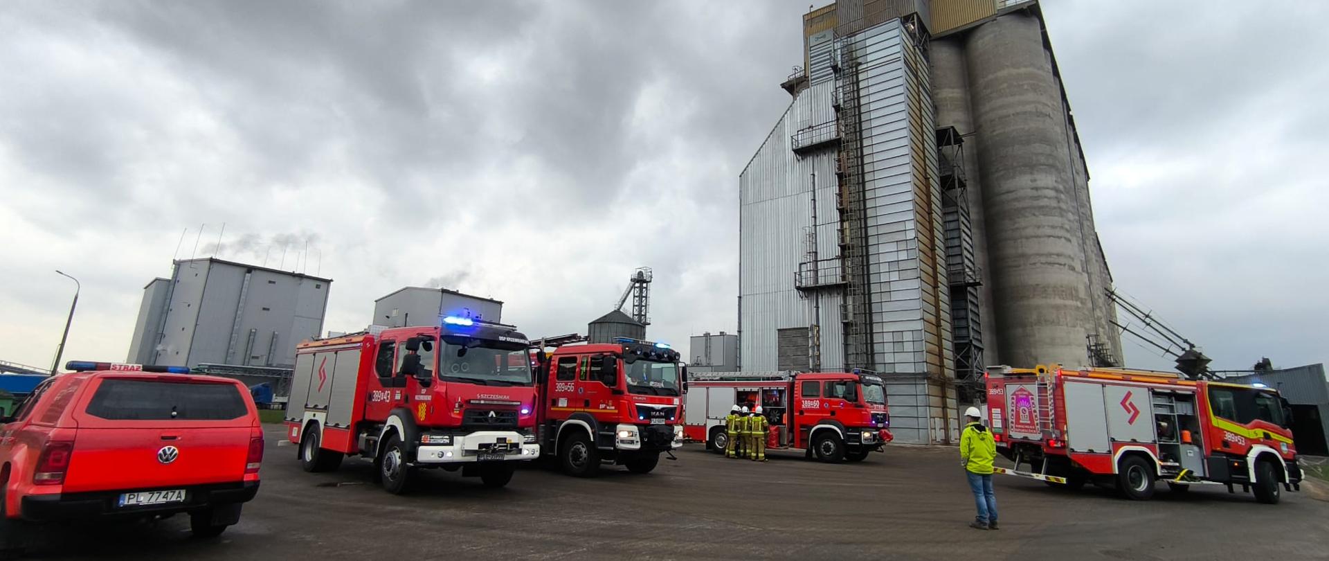 Samochody strażackie ustawione na placu przed silosami zbożowymi.