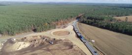 Skrzyżowanie z lotu ptaka na etapie prac ziemnych z widocznym zarysem ronda, wkomponowane u zbiegu dwóch dróg, po których odbywa się ruch samochodów. Dookoła lasy i pola.
