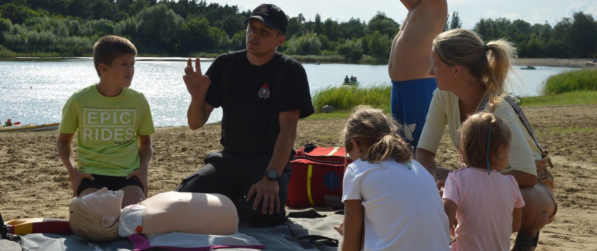 Piknik "Kręci mnie bezpieczeństwo nad wodą". Na kocu manekin do nauki zasad udzielania pierwszej pomocy, a przy nim klęczący strażak, który instruuje w jaki sposób prowadzić zewnętrzny masaż serca i sztuczne oddychanie. Wokół czworo dzieci i kobieta. Jeden z chłopców ma uniesione ręce. W tle akwen.