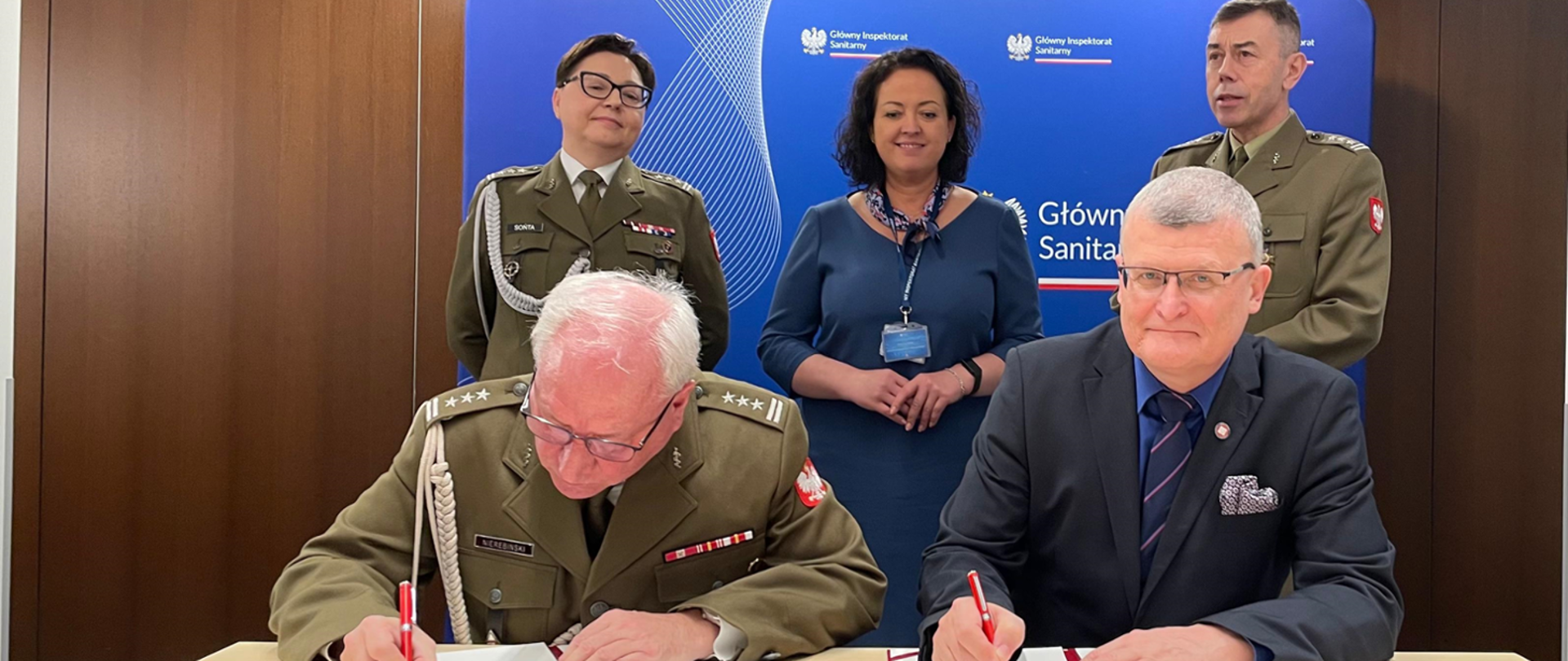 Na zdjęciu widzimy moment podpisania dokumentu przez dwie osoby siedzące przy stole. Jeden z mężczyzn nosi mundur wojskowy z odznaczeniami i pagonami generalskimi, a drugi jest ubrany w ciemny garnitur, koszulę i krawat. Obaj trzymają czerwone długopisy i koncentrują się na podpisywaniu dokumentu.
Za nimi stoją trzy osoby: dwie w mundurach wojskowych oraz jedna kobieta w granatowej sukience i z identyfikatorem na smyczy. W tle widoczna jest tablica z napisem "Główny Inspektorat Sanitarny" oraz polskim godłem, co sugeruje, że wydarzenie ma oficjalny charakter i dotyczy spraw związanych z bezpieczeństwem sanitarnym lub wojskową służbą zdrowia.
Cała scena wygląda na oficjalne spotkanie o dużym znaczeniu, prawdopodobnie dotyczące współpracy między wojskiem a służbami sanitarnymi.